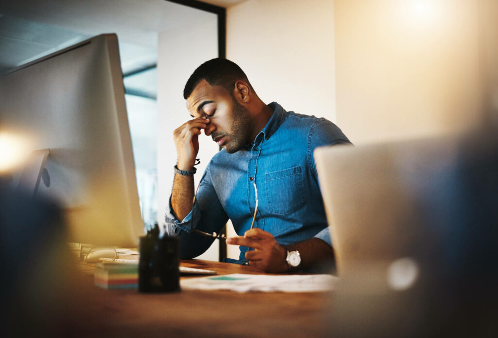 Man pinching bridge of nose in stress while sitting in front of work computer.