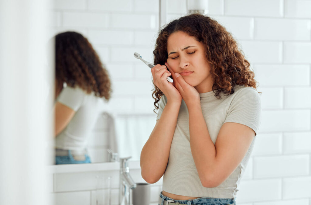 Toothache, pain and sensitive teeth with a woman brushing her teeth in a bathroom at home.